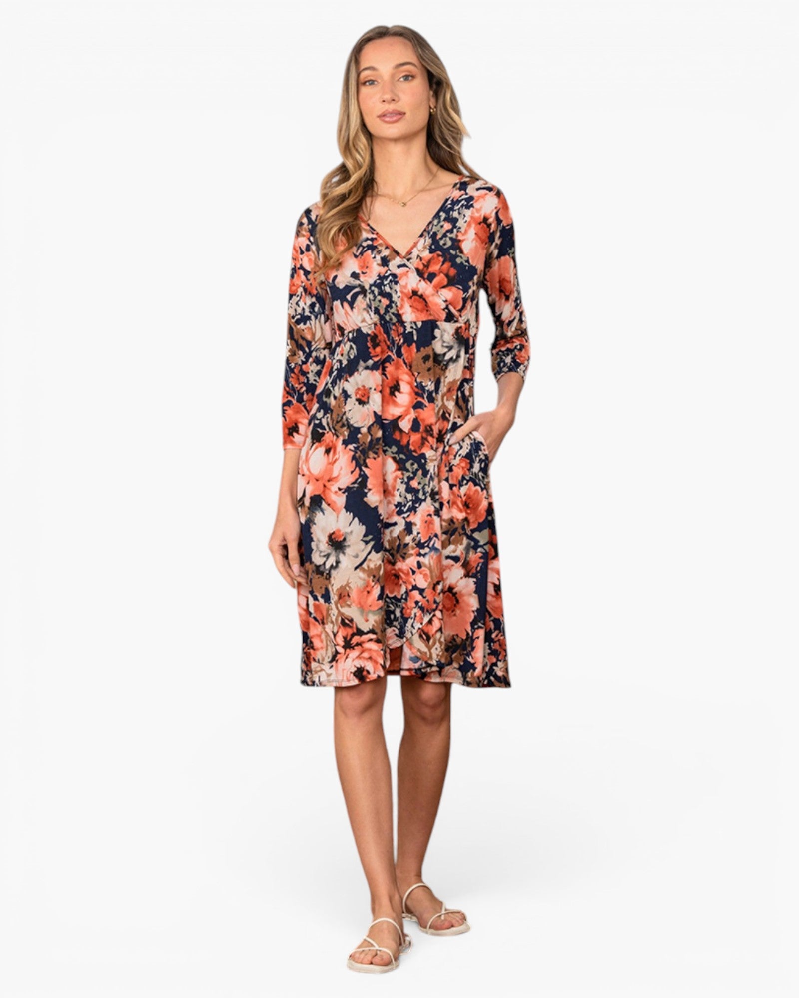A woman modeling a knee-length floral dress with three-quarter sleeves and side pockets, featuring a navy blue base with orange, pink, and white flowers, against a plain white background.