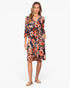 A woman modeling a knee-length floral dress with three-quarter sleeves, a V-neckline, and side pockets, shown against a plain light gray studio background.