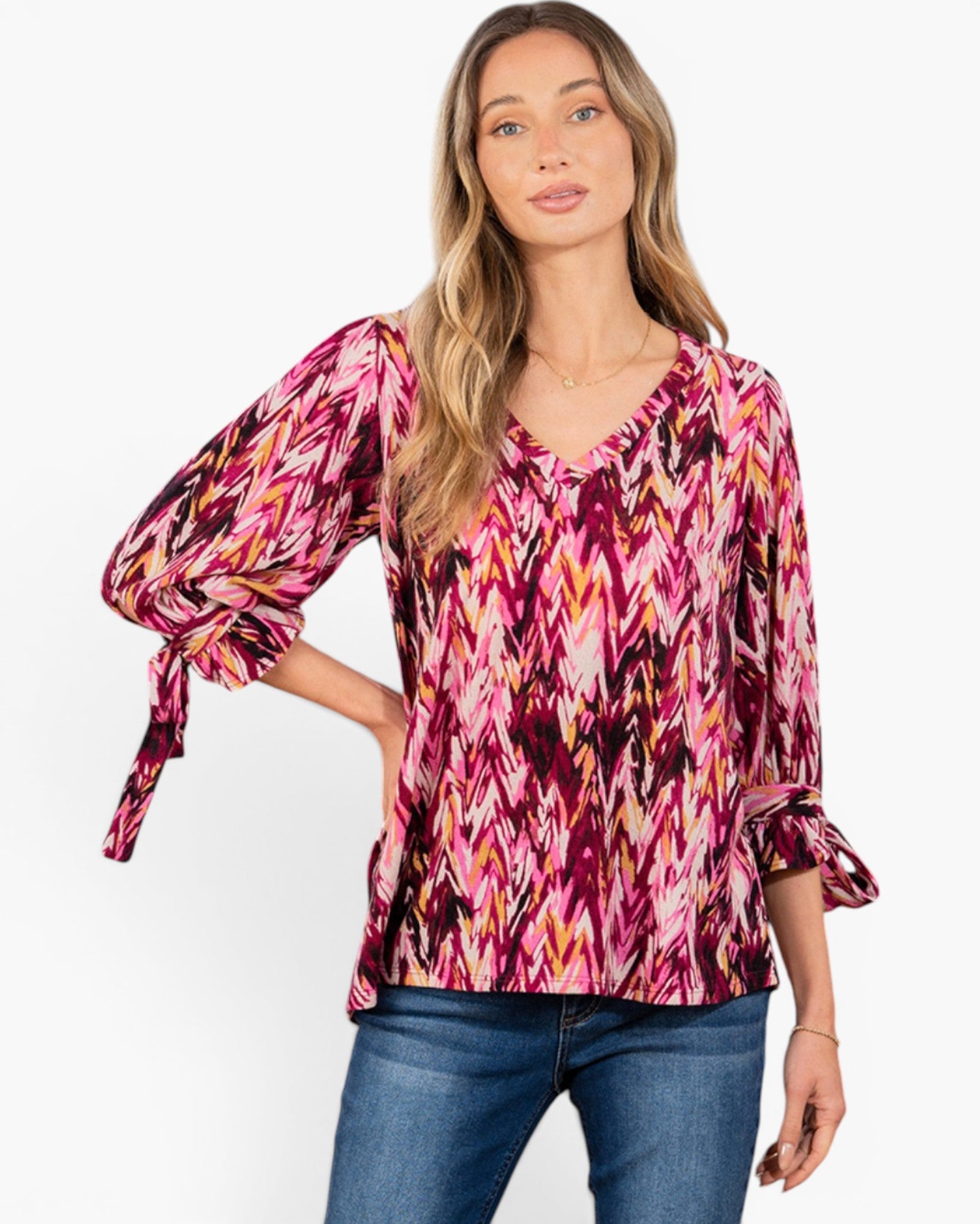 A woman modeling a pink and magenta chevron-patterned blouse with three-quarter sleeves and tie details, paired with blue jeans against a plain white background.