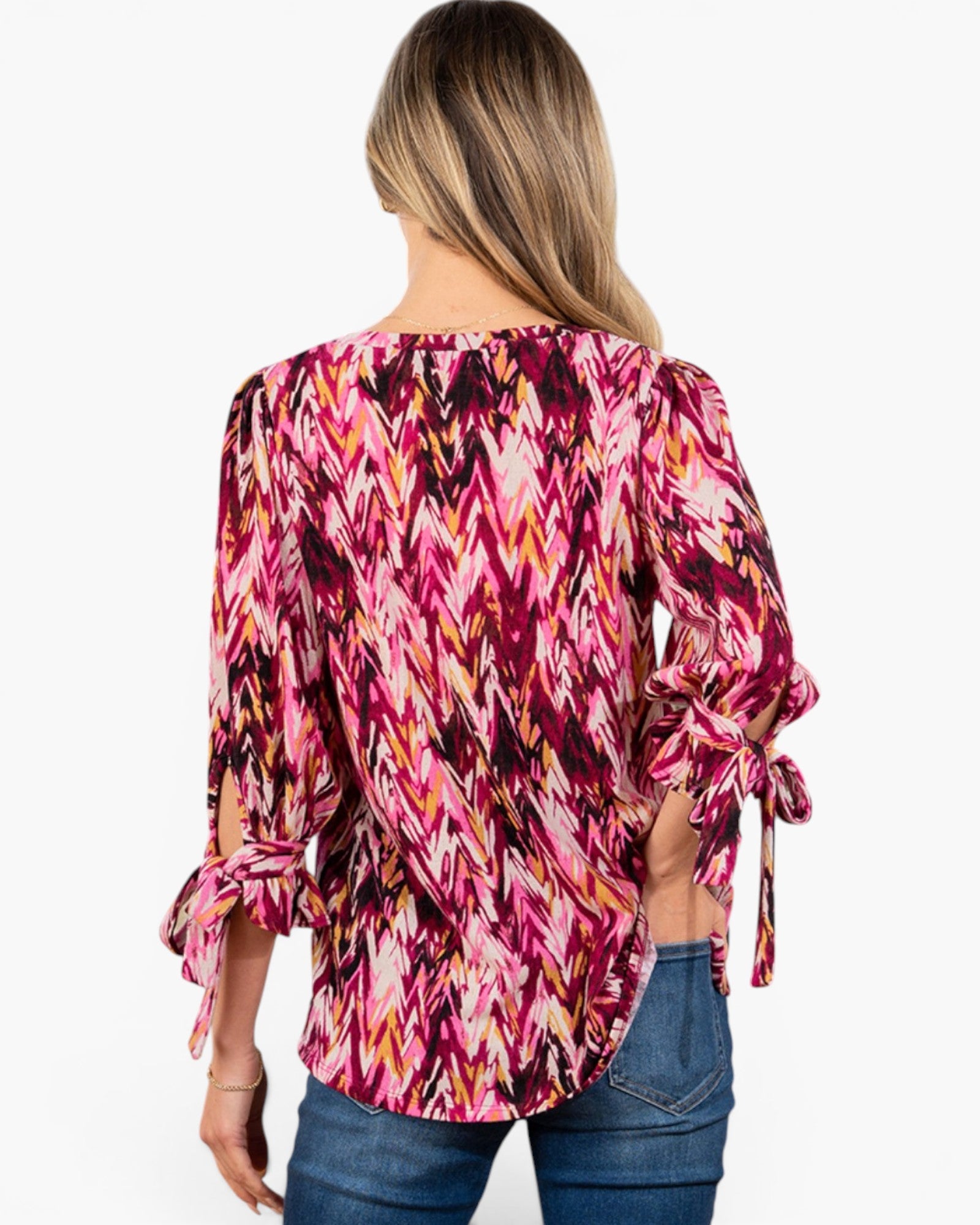 A woman viewed from the back wearing a pink, magenta, and orange chevron-patterned blouse with tie details on the sleeves, paired with blue jeans, against a plain white background.