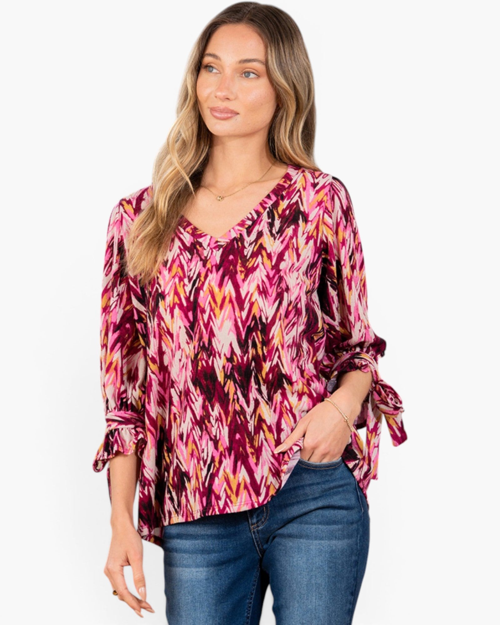 A woman modeling a long-sleeve blouse with a vibrant pink, purple, and orange chevron pattern, paired with blue jeans against a plain white background.