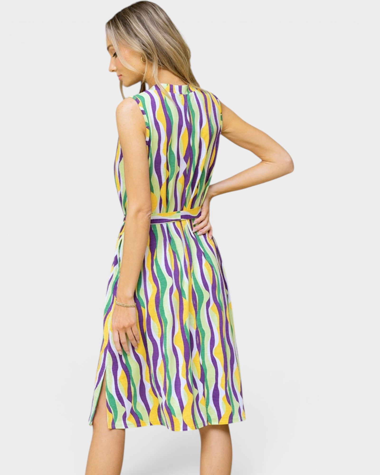 A woman modeling a sleeveless knee-length dress with a colorful abstract wavy pattern in purple, yellow, green, and white, shown from a three-quarter back angle against a light gray studio background.