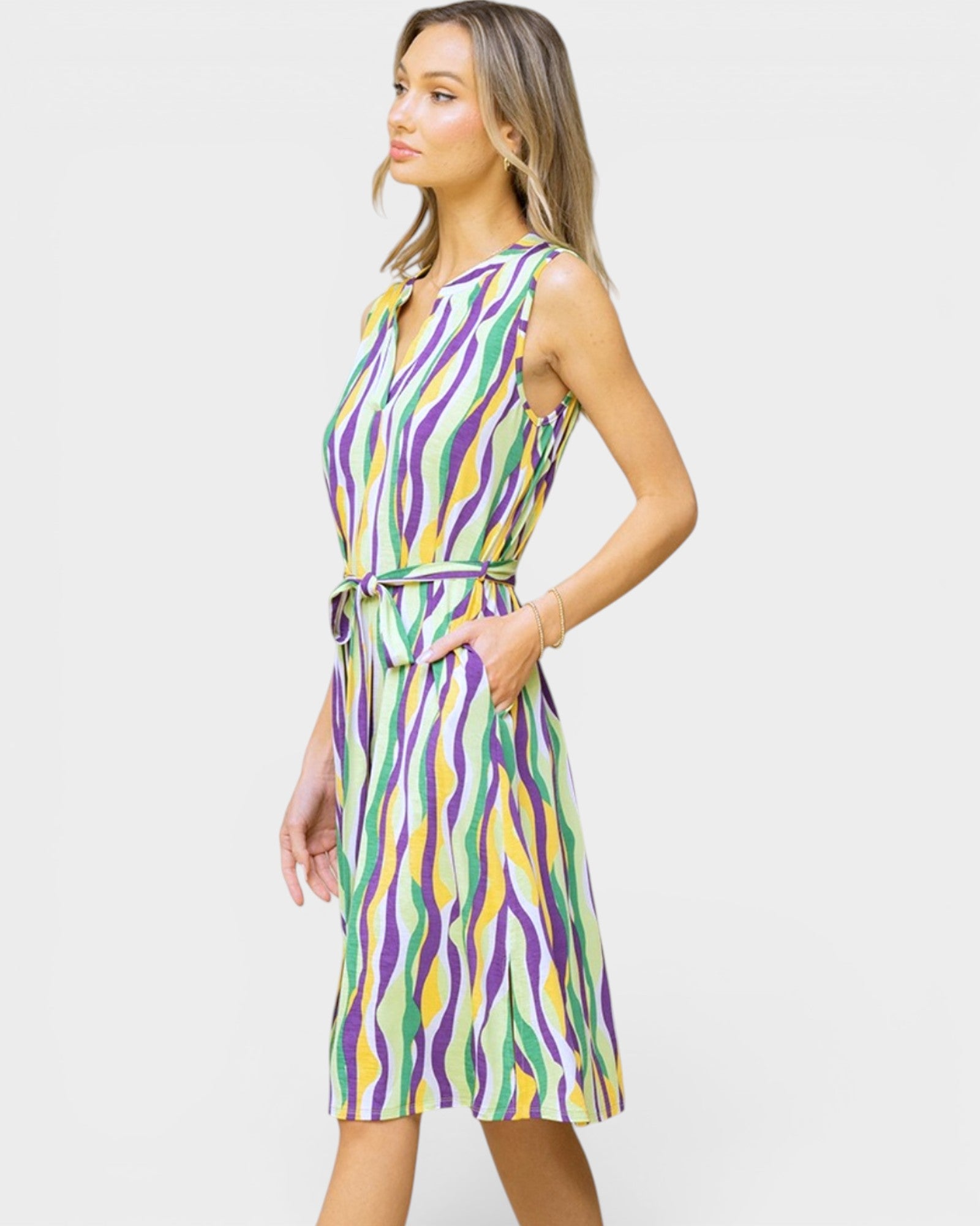 A woman modeling a sleeveless knee-length dress with a colorful abstract wavy stripe pattern in purple, green, yellow, and white, featuring a tie waist belt, set against a plain light gray background.
