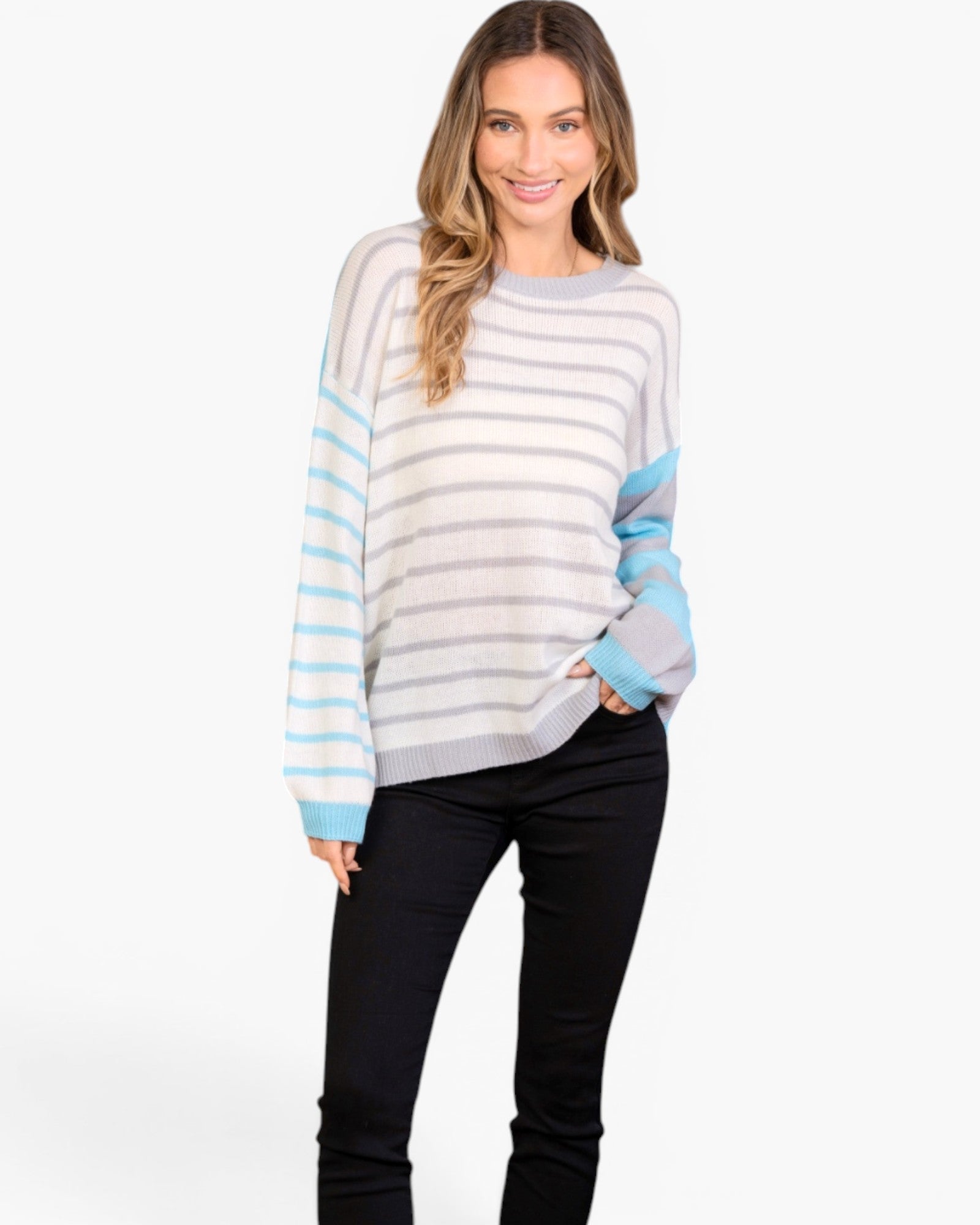 A woman modeling a color-blocked knit sweater with horizontal light gray and white stripes, light blue sleeve panels, and a light gray crew neck, paired with black pants against a plain white studio background.