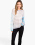 A woman modeling a color-blocked knit sweater with horizontal light gray and white stripes, light blue sleeve panels, and a light gray crew neck, paired with black pants against a plain white studio background.
