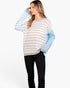 A woman modeling a light gray and white striped knit sweater with light blue color-blocked sleeves, paired with black pants, against a plain white studio background.