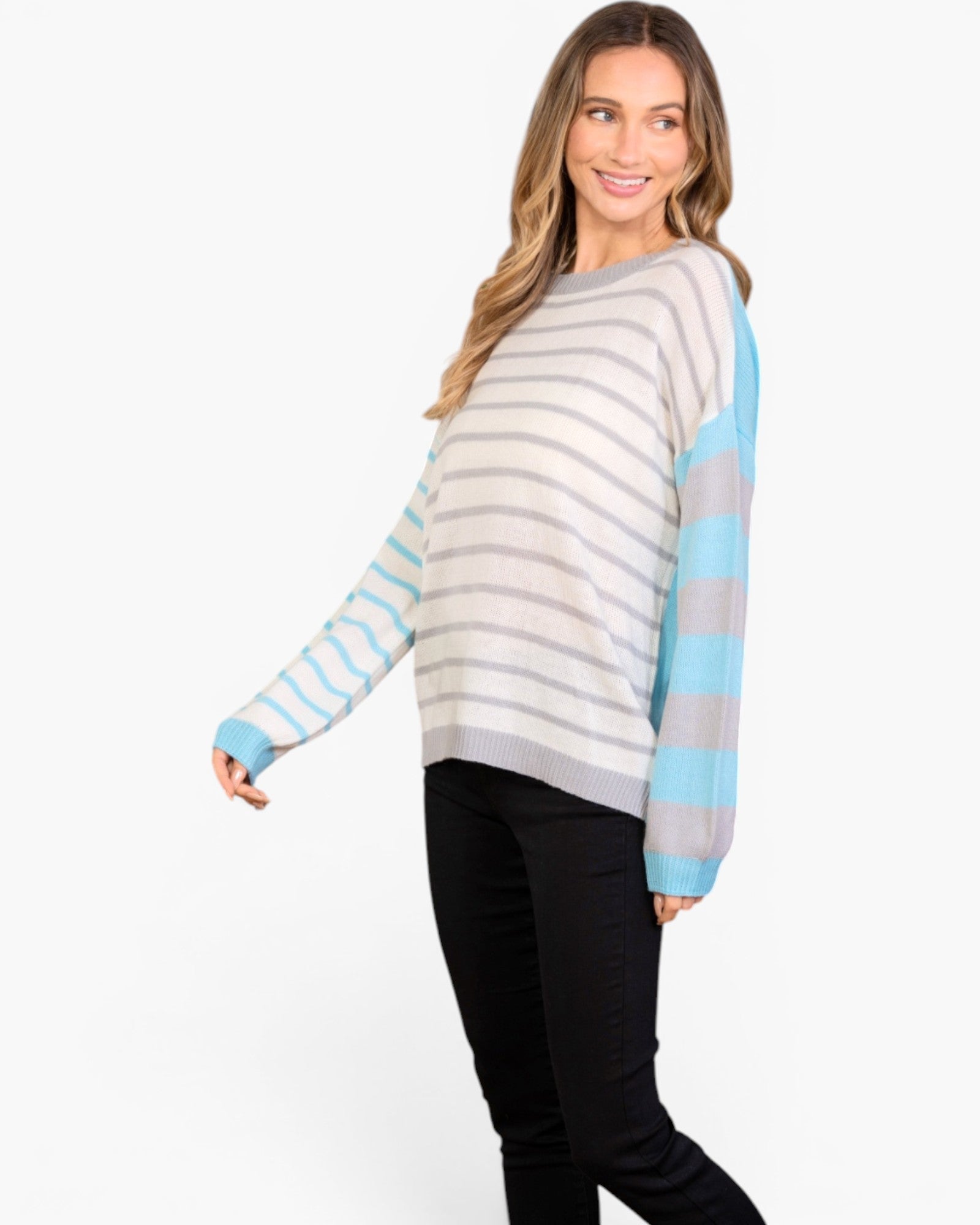 A woman modeling a knit sweater with horizontal gray and white stripes on the body and light blue sleeves, paired with black pants, against a plain white studio background.