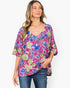A woman wearing a vibrant floral print top with blue, green, and pink flowers on a magenta background, paired with blue jeans, against a plain white studio background.