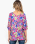 A woman viewed from the back wearing a bright pink floral top with blue and green flowers, paired with blue jeans, against a plain white background.