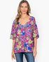 A woman wearing a vibrant floral print top with blue, green, and pink flowers, paired with blue jeans, against a plain white background.