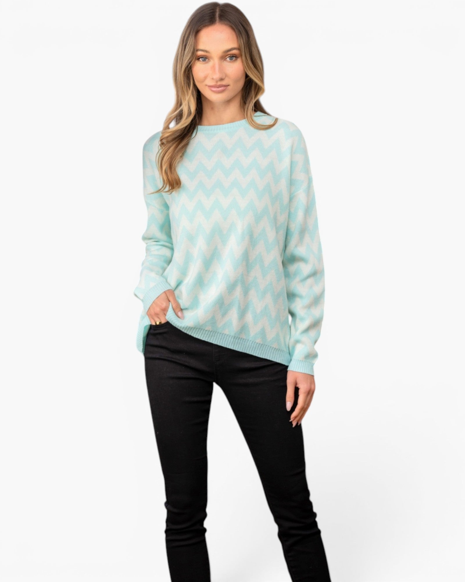 A woman modeling a mint green and white chevron-patterned knit sweater with black pants, standing against a plain white studio background.