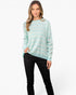 A woman modeling a mint green and white chevron-patterned knit sweater with black pants, standing against a plain white studio background.