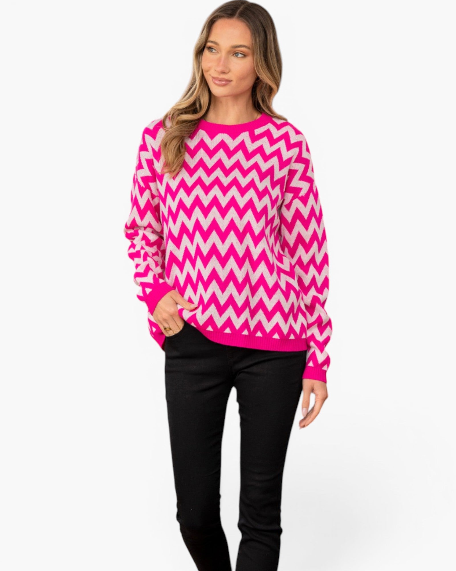 A woman modeling a bright pink and white chevron patterned knit sweater with black pants, photographed against a clean white studio background.