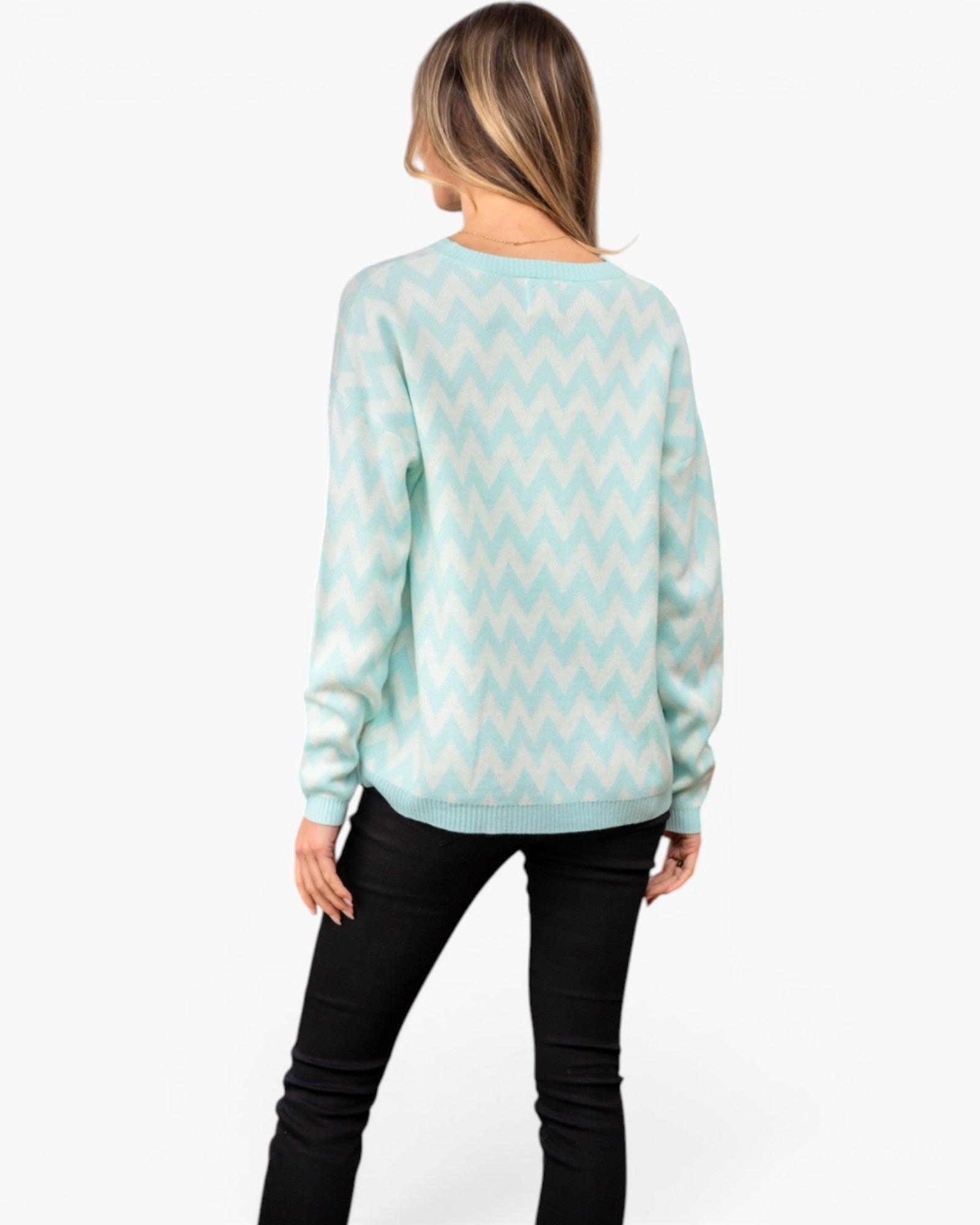 A woman viewed from the back wearing a mint green and white chevron knit sweater and black pants, set against a plain white studio background.