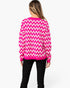 A woman viewed from the back wearing a bright pink and white chevron patterned sweater and black pants, against a plain light gray studio background.