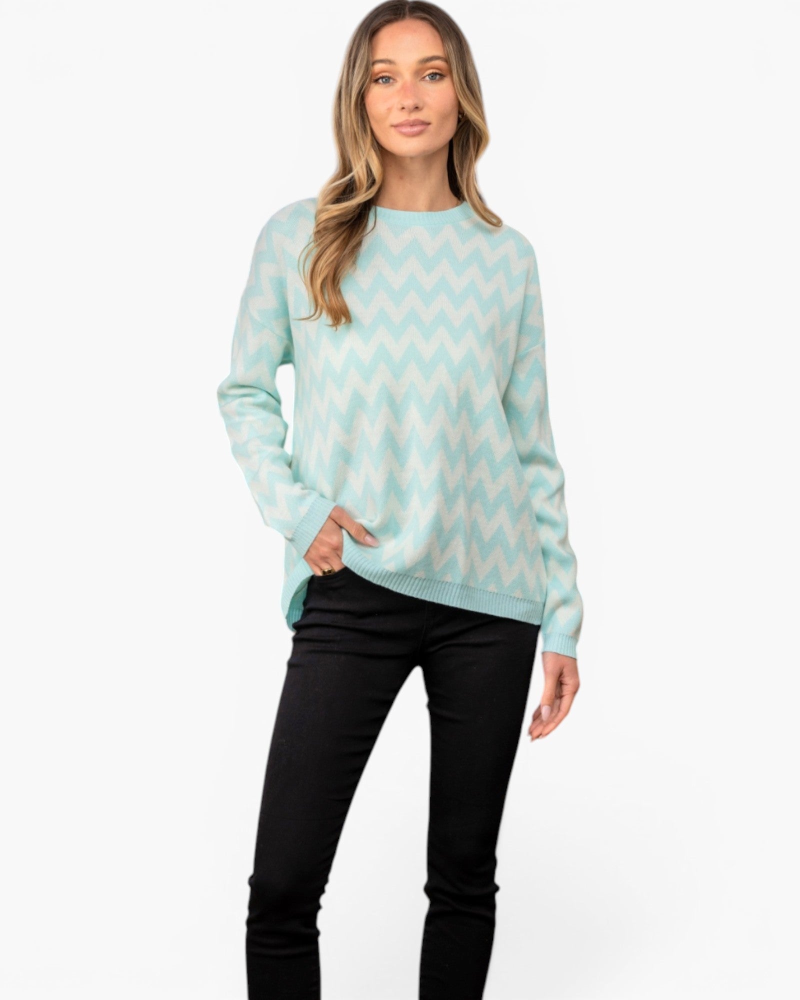 A woman modeling a mint green and white chevron-patterned knit sweater with black pants, standing against a plain white studio background.