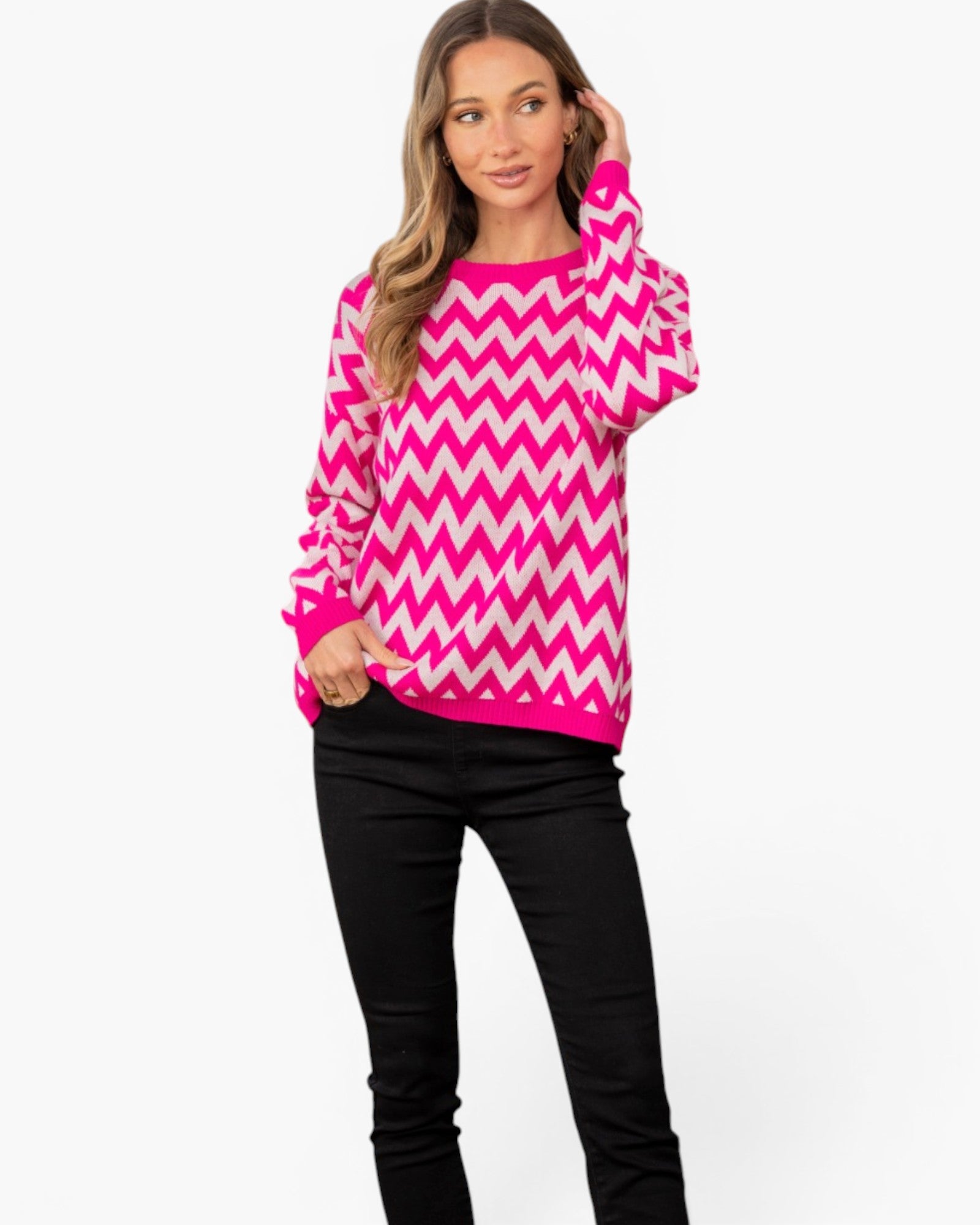 A woman modeling a bright pink and white chevron-patterned knit sweater with black pants, standing against a plain white studio background.