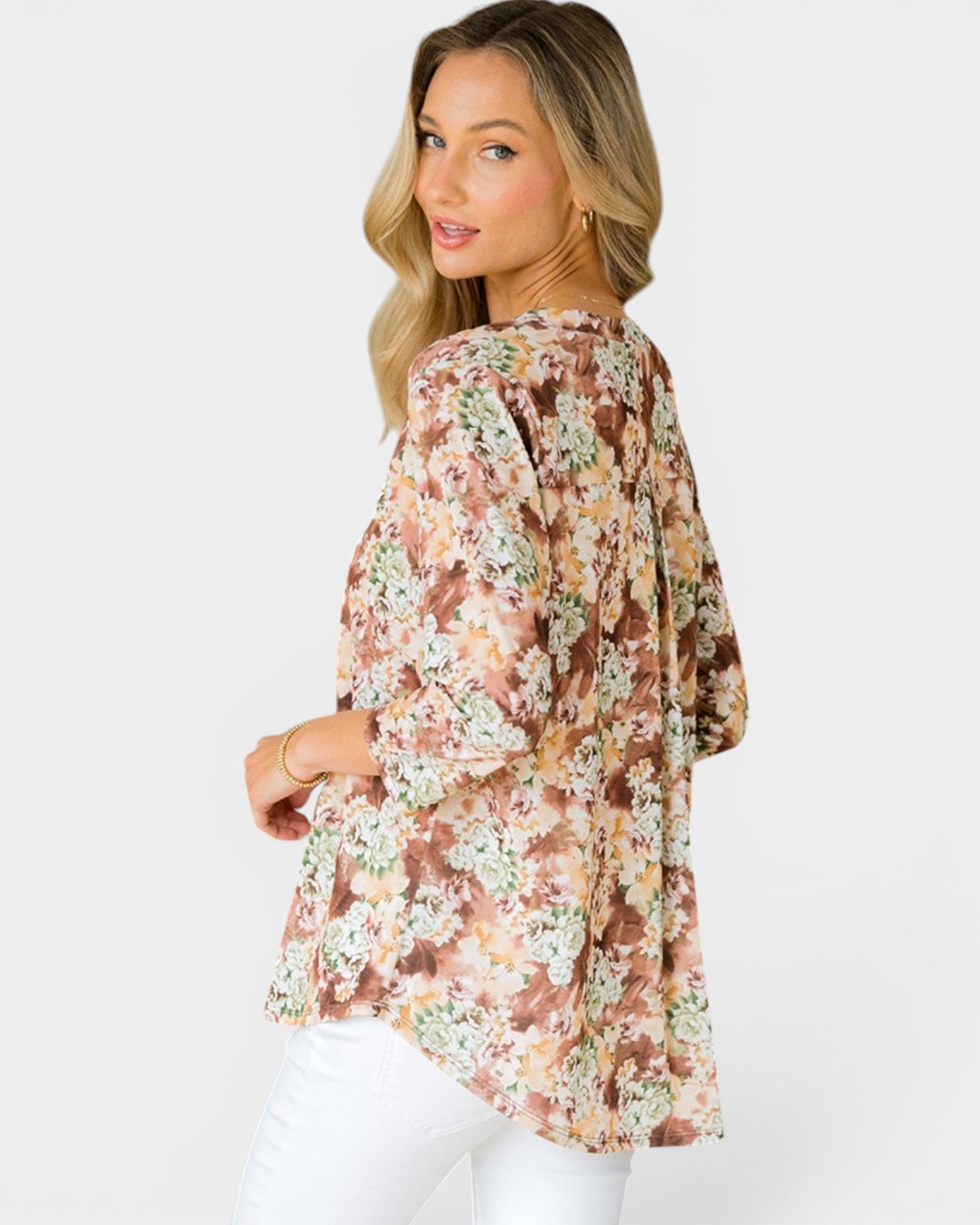 A woman wearing a loose, three-quarter sleeve floral top with a brown, beige, and white pattern, paired with white pants, photographed against a light gray studio background.