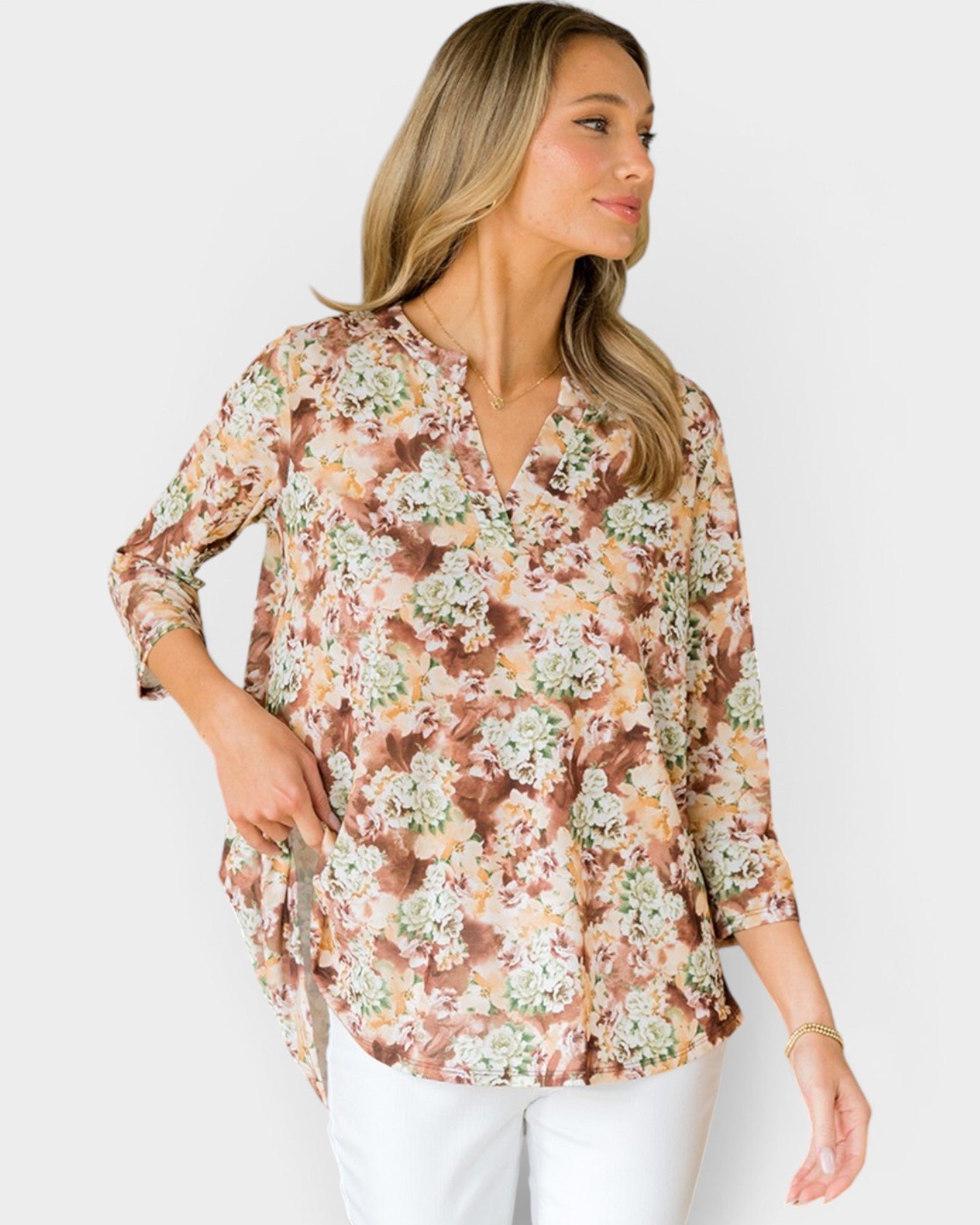 A woman wearing a three-quarter sleeve button-up blouse with a brown, beige, and white floral pattern, paired with white pants, photographed against a light gray studio background.