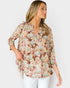 A woman wearing a three-quarter sleeve button-up blouse with a brown, beige, and white floral pattern, paired with white pants, photographed against a light gray studio background.