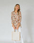 Woman wearing a floral blouse and holding a tote bag against a plain background