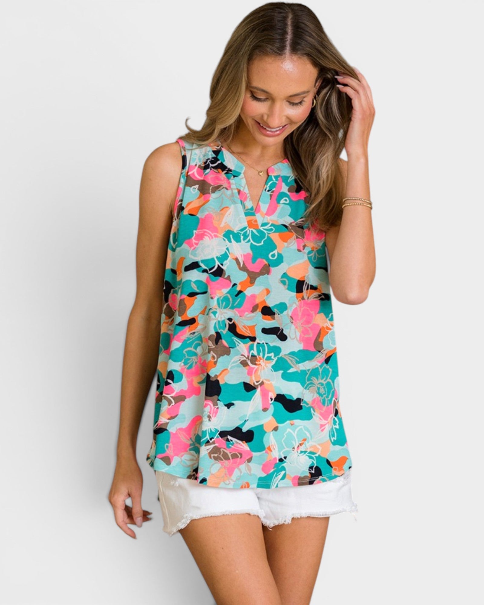 A woman wearing a sleeveless, V-neck top with a colorful abstract print in turquoise, pink, orange, black, and white, paired with white denim shorts, set against a plain light gray background.