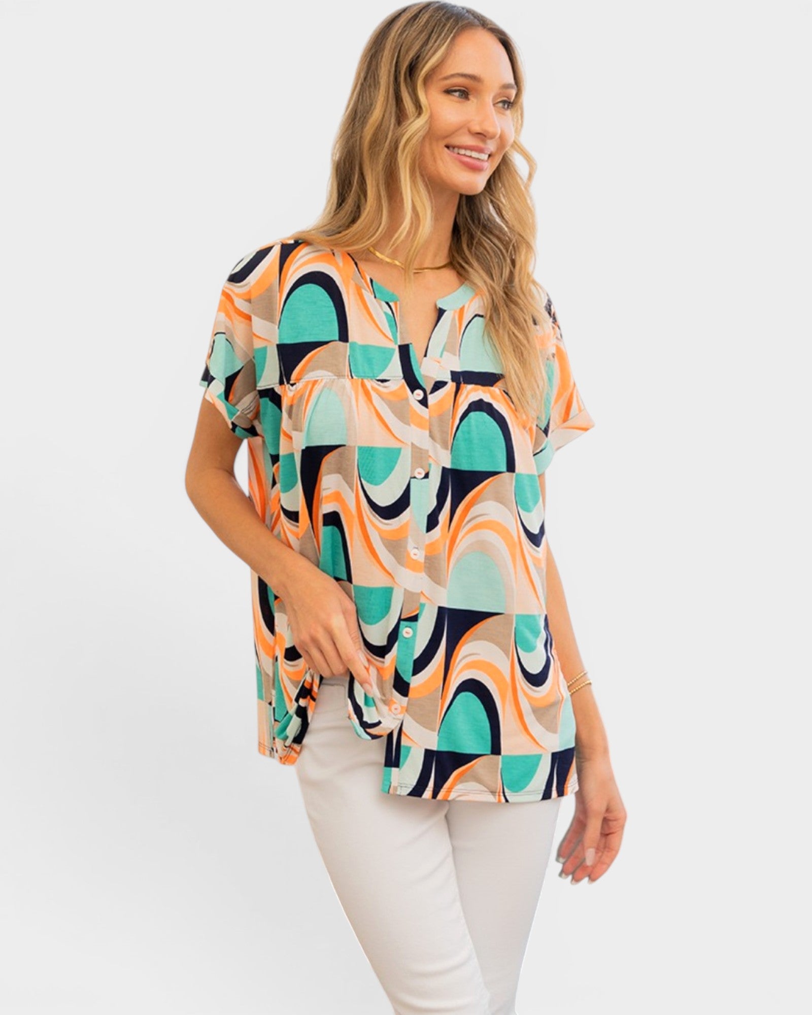 A woman modeling a short-sleeve button-front blouse with an abstract geometric print in teal, orange, navy, and beige, paired with white pants against a light gray studio background.