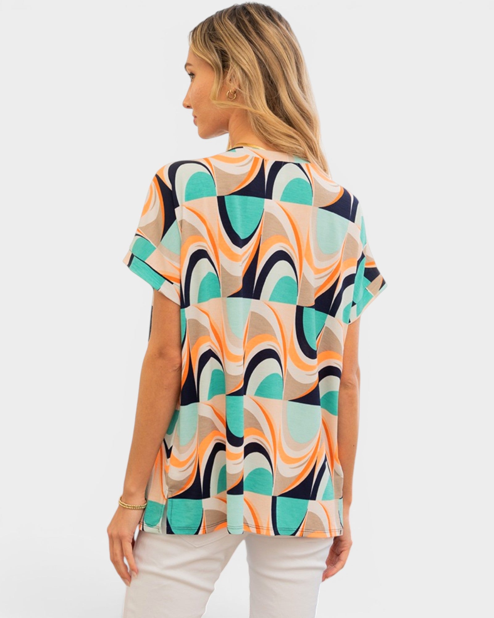 A woman shown from the back wearing a short-sleeve top with a bold abstract geometric pattern in teal, orange, beige, and black, paired with white pants, against a light gray studio background.