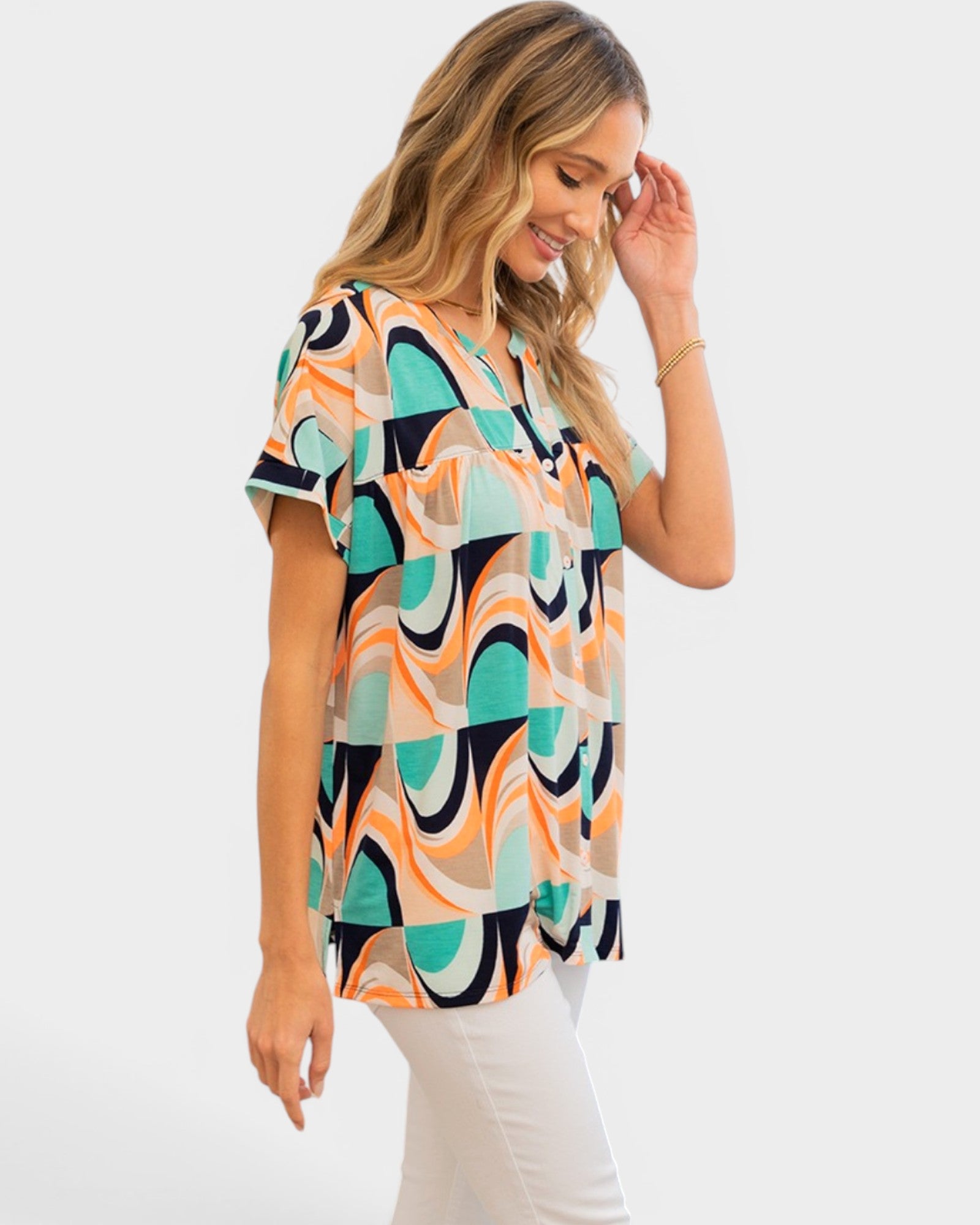 A woman wearing a short-sleeve V-neck top with a colorful abstract geometric pattern in teal, orange, black, and cream, paired with white pants, photographed against a light gray studio background.