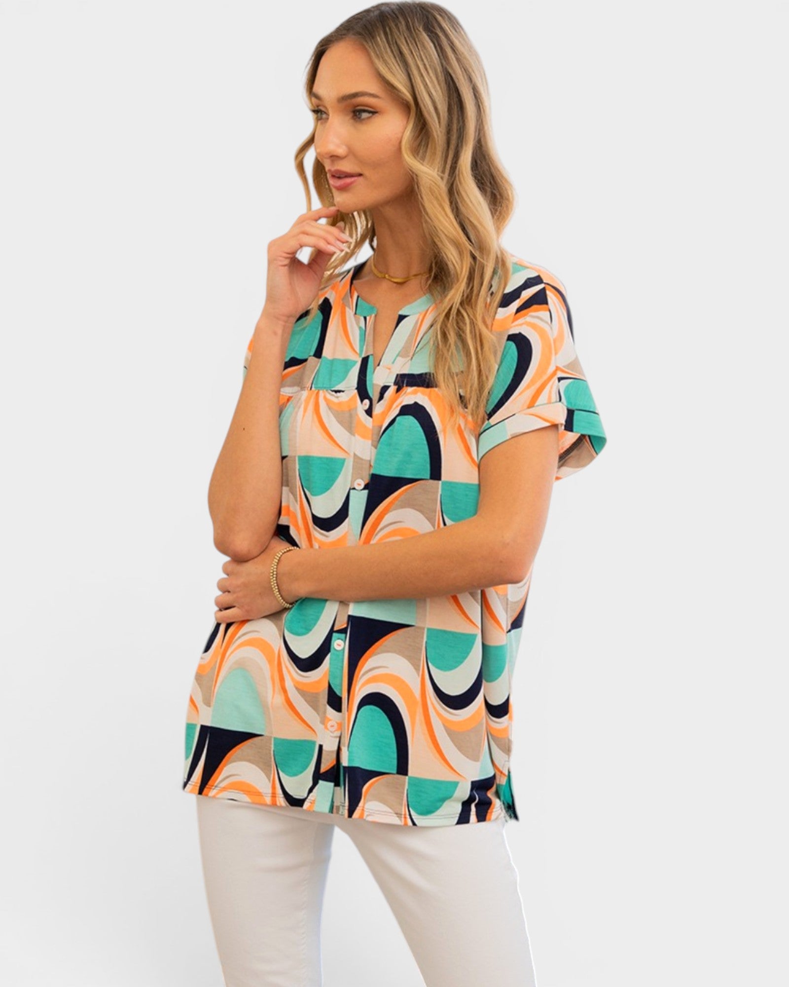 A woman modeling a short-sleeve button-up blouse with a colorful abstract geometric pattern in teal, orange, black, and beige, paired with white pants against a light gray studio background.