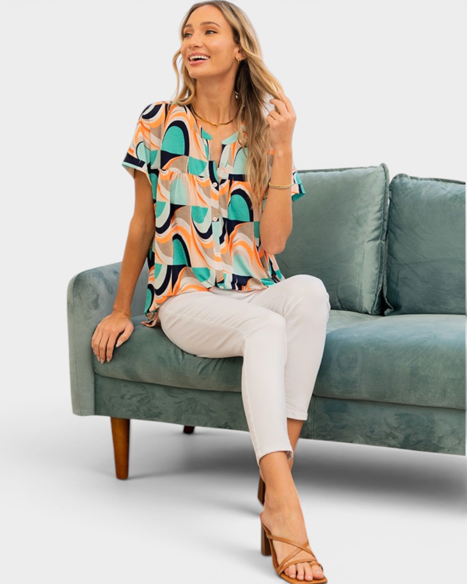 A woman models a short-sleeve blouse with a colorful abstract geometric pattern in teal, peach, black, and white, paired with white pants and tan heeled sandals, sitting on a teal velvet sofa against a light gray background.