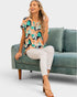 A woman models a short-sleeve blouse with a colorful abstract geometric pattern in teal, peach, black, and white, paired with white pants and tan heeled sandals, sitting on a teal velvet sofa against a light gray background.