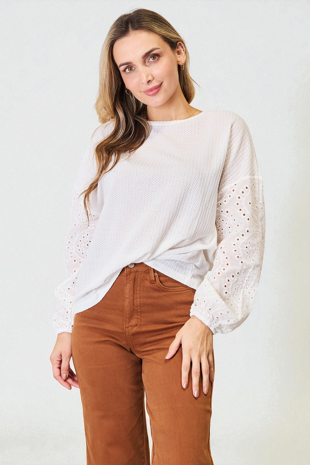 Woman wearing a white sweater with lace sleeves and brown pants on a light background