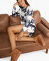 A woman sits on a brown leather couch wearing a long-sleeve tie-dye mini dress with a blue and white cloud pattern, paired with brown suede platform boots.