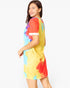 A woman modeling a short-sleeve tie-dye t-shirt dress with yellow, red, blue, and purple colors, shown from a side-back angle against a plain white background.