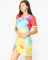 A woman modeling a short-sleeve V-neck tunic dress with a vibrant tie-dye pattern in yellow, blue, red, and purple, featuring white trim on the sleeve cuffs, set against a plain white background.