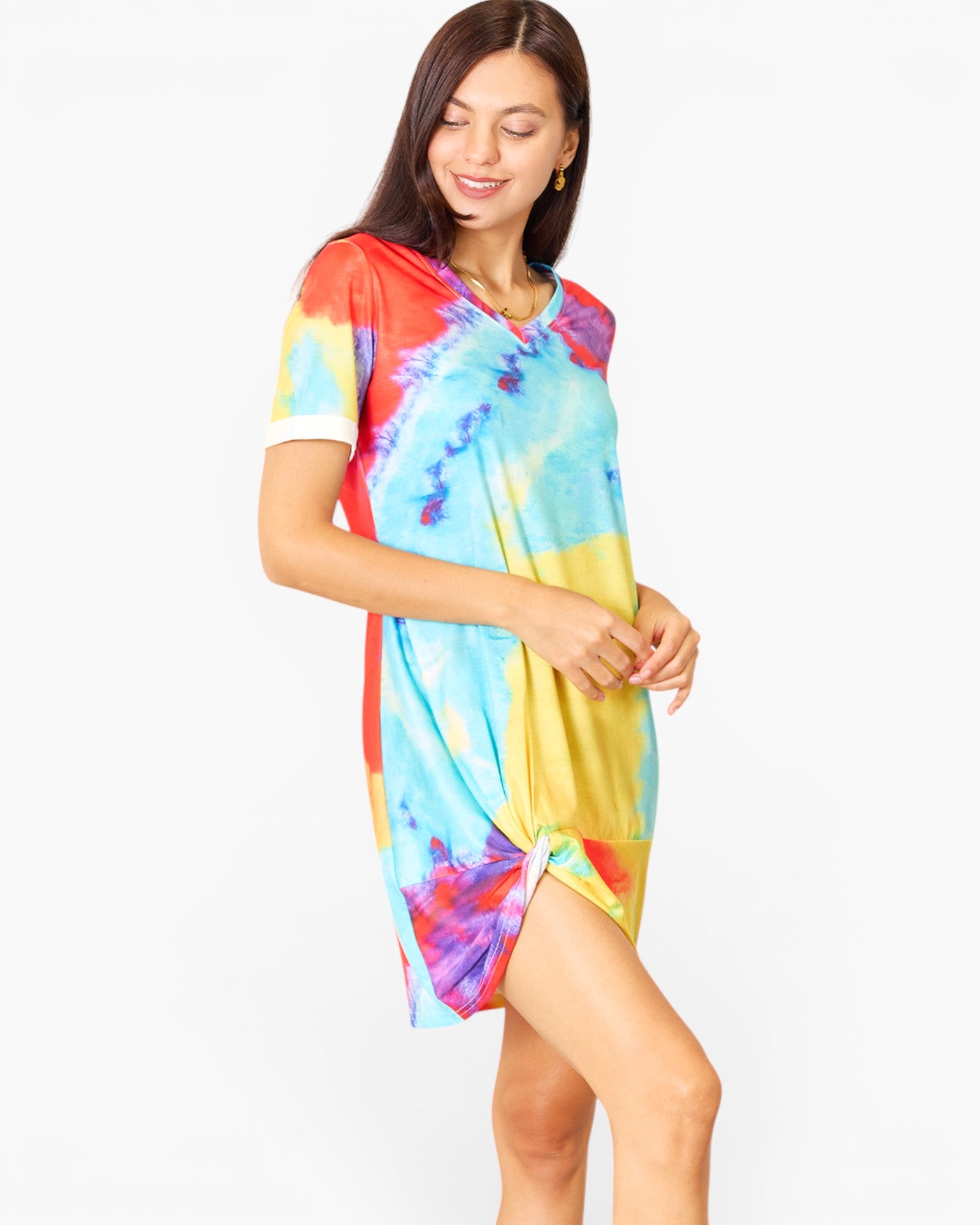 A woman modeling a short-sleeve tie-dye tunic dress with a vibrant mix of blue, yellow, red, and purple colors, set against a plain white studio background.