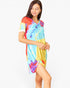 A woman modeling a short-sleeve tie-dye tunic dress with a vibrant mix of blue, yellow, red, and purple colors, set against a plain white studio background.