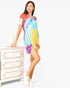 A woman models a short-sleeve tie-dye t-shirt dress with a vibrant multicolor pattern of blue, yellow, orange, and purple. She is standing next to a white three-drawer dresser against a plain white studio background.