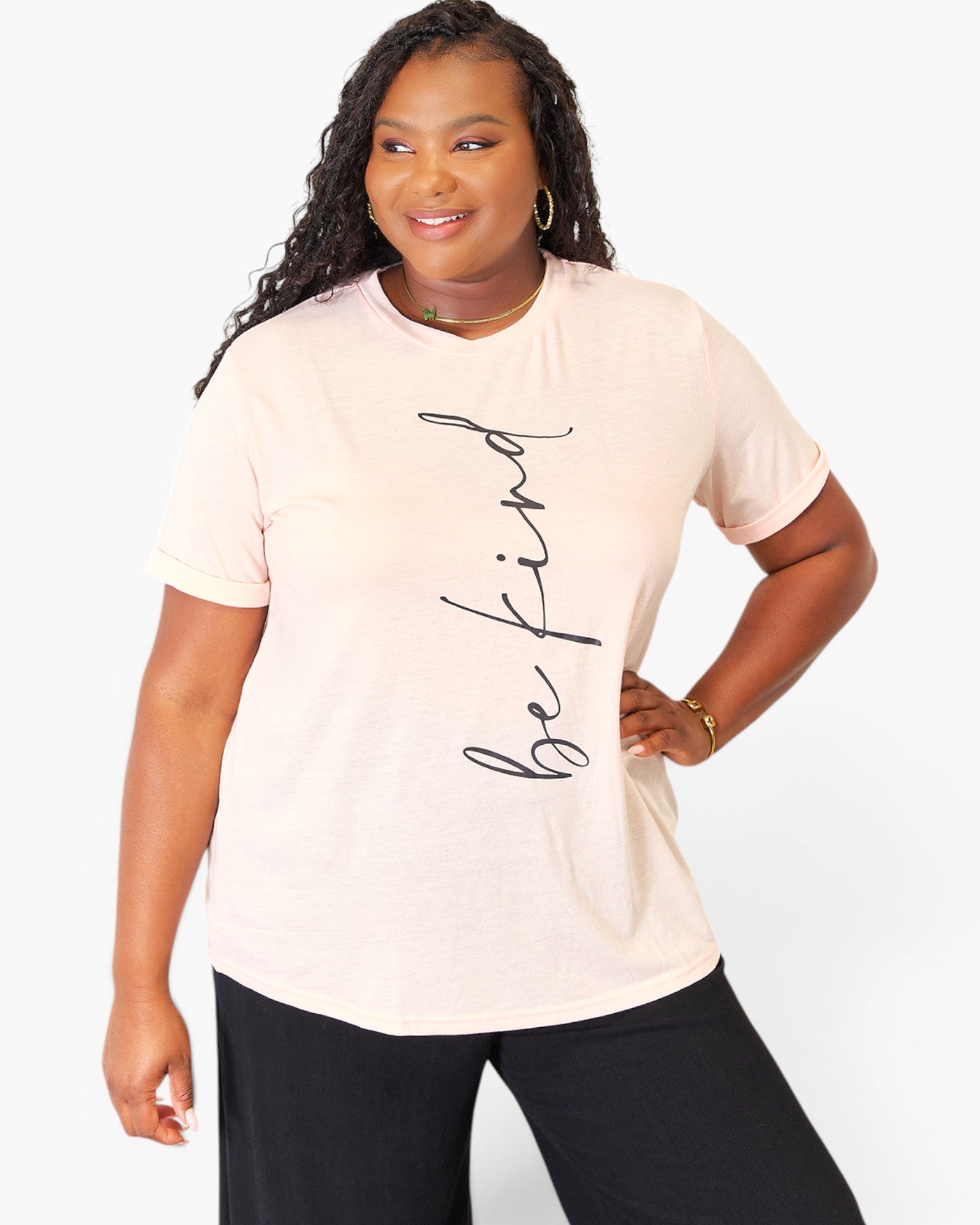 A woman modeling a light peach-pink short-sleeve t-shirt with the phrase 'be kind' printed vertically in black script on the front, paired with black pants, against a plain white studio background.