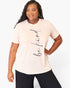 A woman modeling a light peach-pink short-sleeve t-shirt with the phrase 'be kind' printed vertically in black script on the front, paired with black pants, against a plain white studio background.