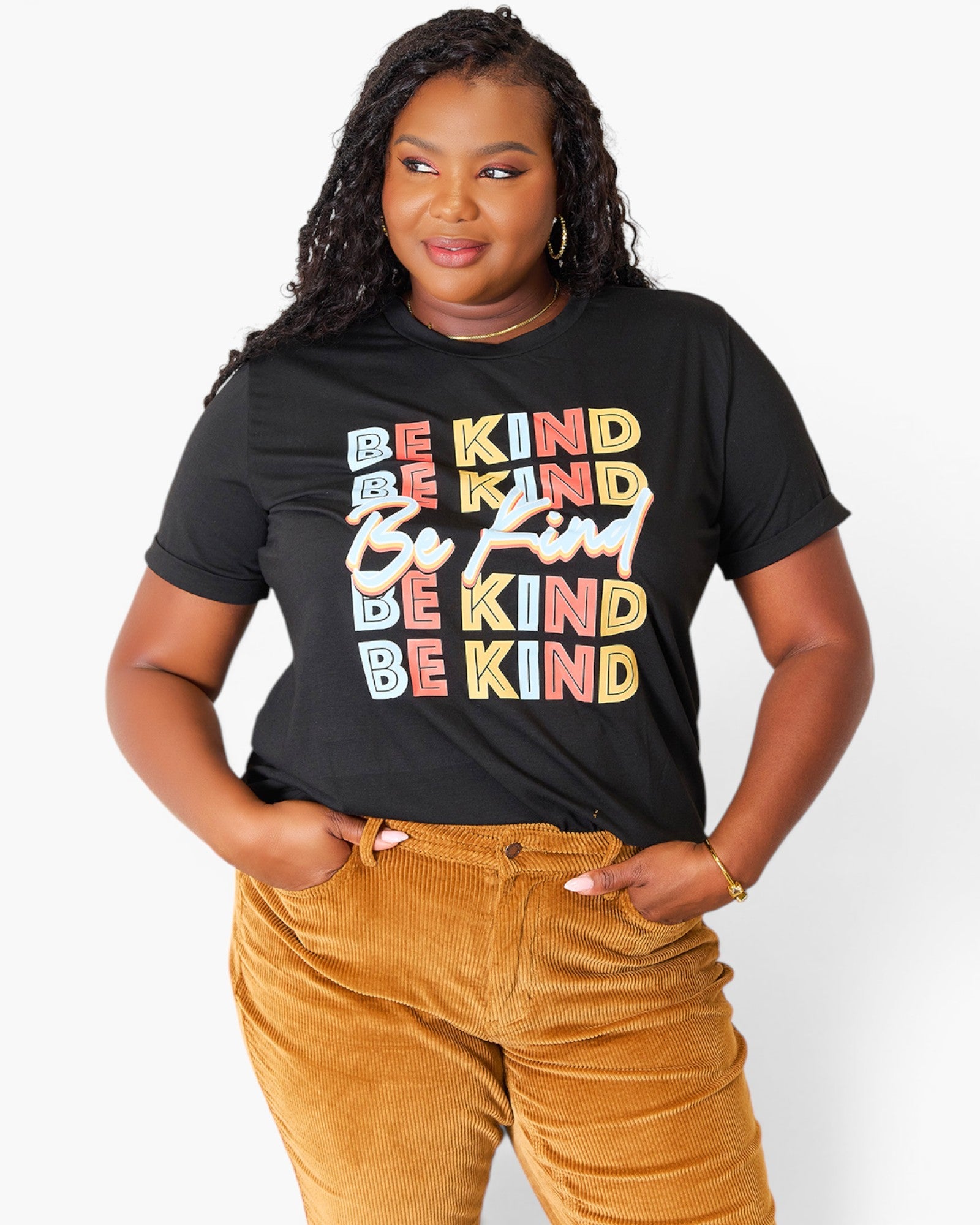 A woman wearing a black graphic t-shirt with the phrase 'BE KIND' repeated in colorful block letters, paired with mustard corduroy pants, against a plain light gray background.