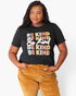 A woman wearing a black graphic t-shirt with the phrase 'BE KIND' repeated in colorful block letters, paired with mustard corduroy pants, against a plain light gray background.