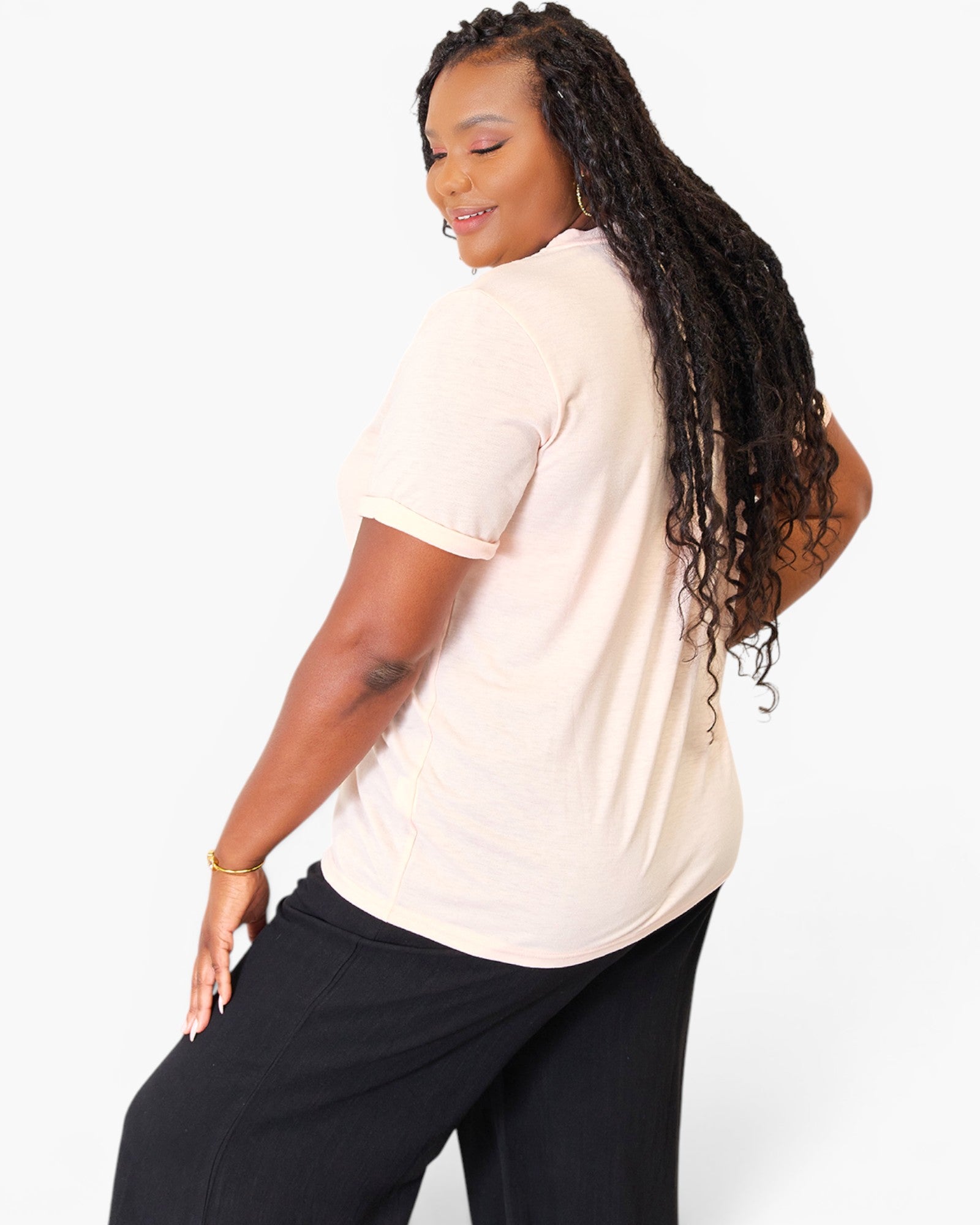 A woman with long, curly black hair is shown from a side-back angle wearing a pale peach short-sleeve t-shirt and black pants against a plain white studio background.