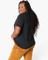 A woman with long, curly black hair is shown from a back-three-quarter angle, wearing a black short-sleeve t-shirt and mustard yellow corduroy pants against a plain white studio background.