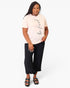 A woman modeling a light pink t-shirt with 'be fired' printed in black script, paired with black cropped pants and black sandals, against a plain white background.