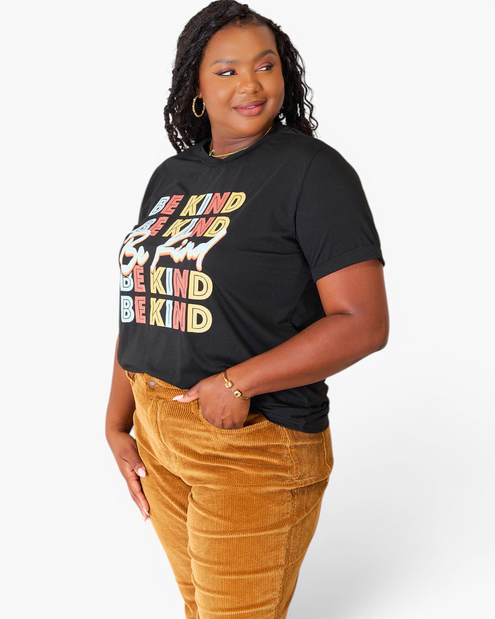 A woman modeling a black graphic t-shirt with a colorful 'BE KIND BE KIND BE KIND' print, paired with mustard yellow corduroy pants, against a plain white studio background.