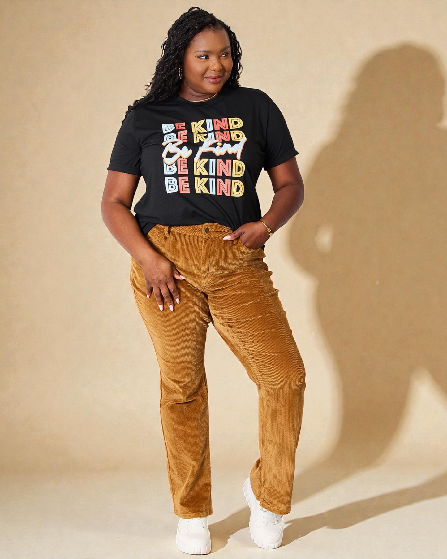 Person wearing a black t-shirt with 'BE KIND' text and brown pants on a beige background