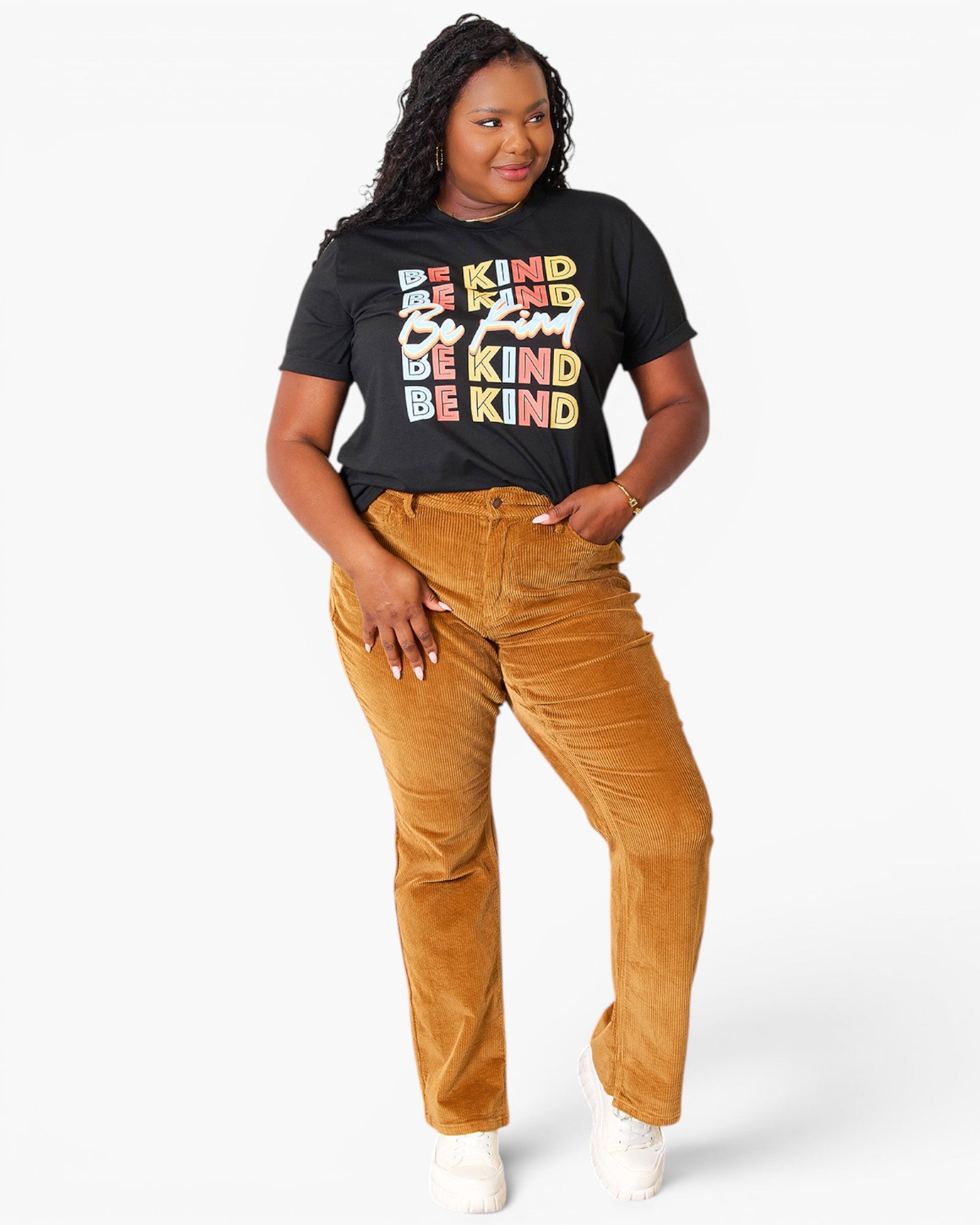 A woman modeling a black graphic t-shirt with the text 'BE KIND' in a multicolor stacked design, paired with mustard yellow corduroy pants and white sneakers, against a plain white background.