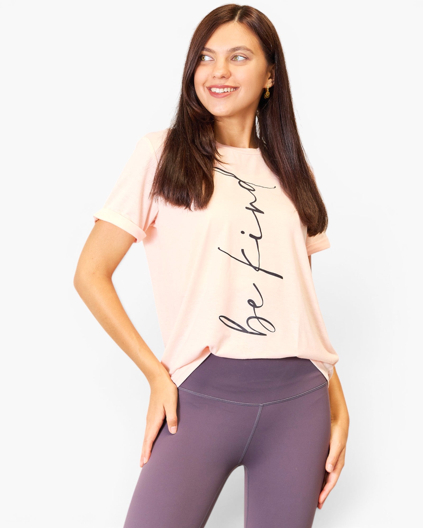A woman wearing a light pink short-sleeve t-shirt with 'be kind' printed in black script, paired with purple leggings, against a light gray background.